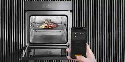 A built-in oven with food visible inside, and a hand holding a smartphone with the control display.