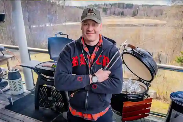 A smiling man stands on a terrace surrounded by grilling tools, with a landscape in the background. On the left is a Weber grill, on the right a black ceramic grill.