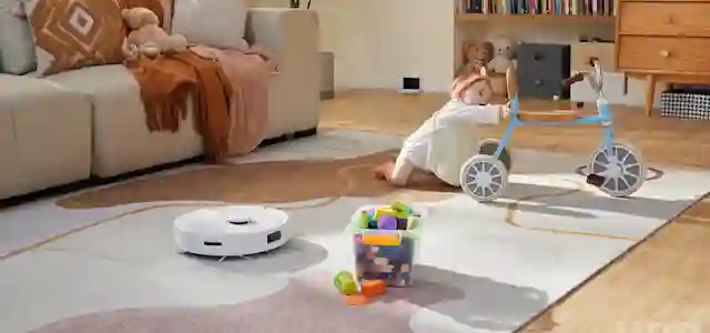 Robotti-imuri imuroi olohuoneessa. jossa pieni lapsi leikkii kolmipyöräisellä.