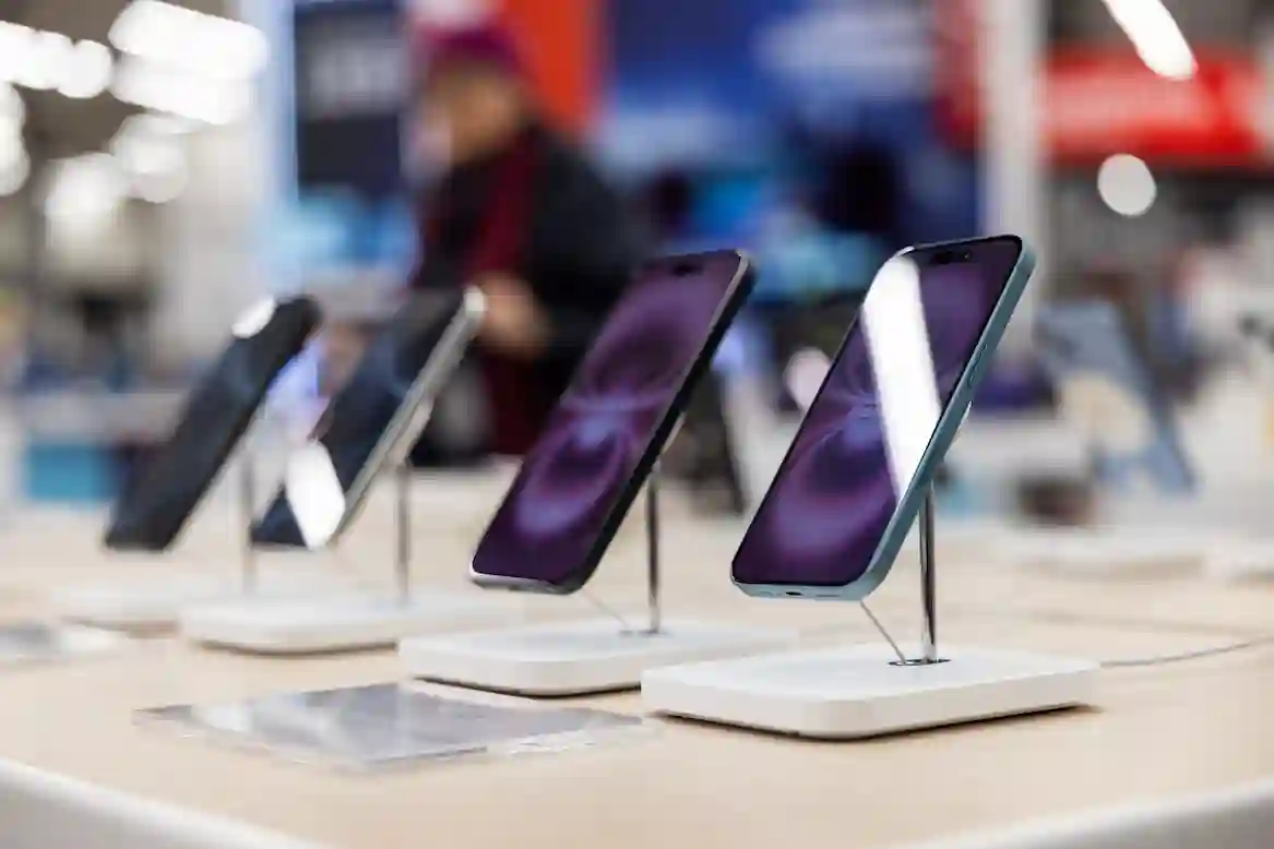 Iphone-puhelimia esillä Helsingin myymälässä puisella esittelypöydällä. Näytöissä on heijastuksia ja taustalla näkyy sumea hahmo.