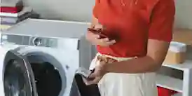 En person i röd tröja använder en smartphone bredvid en tvättmaskin i tvättrummet.