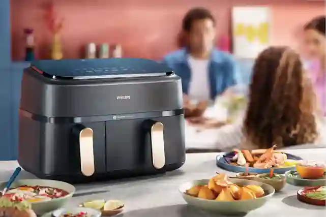 Airfryer keittön tasolla. Taustalla henkilöitä nauttimassa valmiista ateriastat.