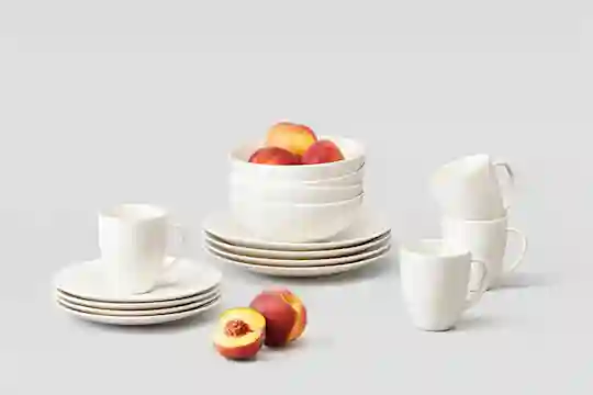 Valkoinen, pelkistetty astiasarja. Lautasia, kulhoja ja kahvikuppeja. Kulhossa persikoita.