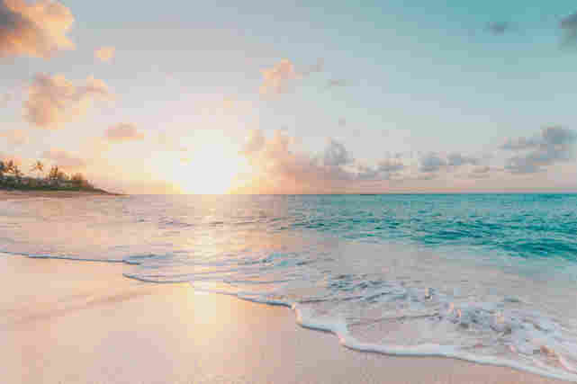 Auringonlasku hiekkarannalla. Aurinko kimaltelee sinisestä vedestä.