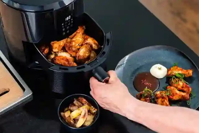 Ströme Compact Air Fryer -airfryer käytössä keittiössä. Siivet ovat valmiita ja henkilö siirtää ne seuraavaksi lautaselleen jossa on kastikkeita ja vihanneksia.