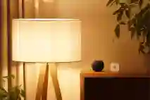 Design valaisin, jonka polttimona toimii lämpimälle värisävylle asetettu älyvalaisin. Vieressä puinen lipasto, jonka päällä Apple HomePod mini sekä liiketunnistin