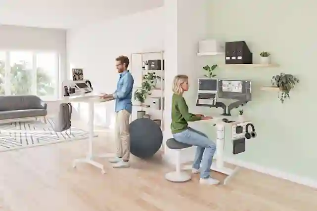 Kotitoimistolla mies tekemässä töitä seisomapöydän ääressä, jonka vieressä jumppapallo. Vieressä toisen työpöydän ääressä istuu nainen ergonomisella työtuolilla.