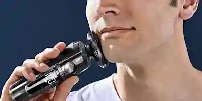 A man shaves his beard with a black electric shaver that has a round shaving head and shaving foam on his face.