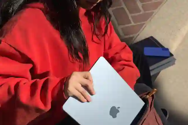 Punapaitainen henkilö työntää kannettavan tietokoneen nahkalaukkuun. Henkilöllä on pitkät mustat hiukset ja harmaassa tietokoneessa on Apple-logo.