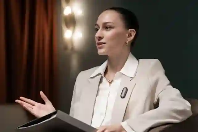 A woman in a beige suit and white shirt sits at a laptop in an interior space with brown curtains and lighting.