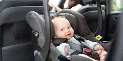 A smiling baby in a gray car seat in the back of a car, with a parent in the driver's seat.
