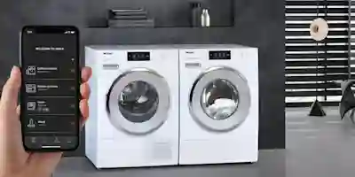 Två vita Miele-torktumlare bredvid varandra i ett modernt badrum, med en smartphone i handen som visar Miele-appen.