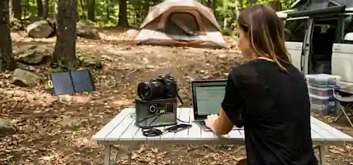 Henkilö käyttää kannettavaa tietokonetta ulkona pöydällä, jossa on myös kamera ja kannettava virtalähde kytkettynä laitteisiin.