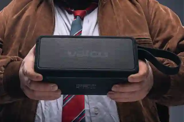 A person holds a black portable Valco Bluetooth speaker in their hands, wearing a brown jacket over a white shirt.