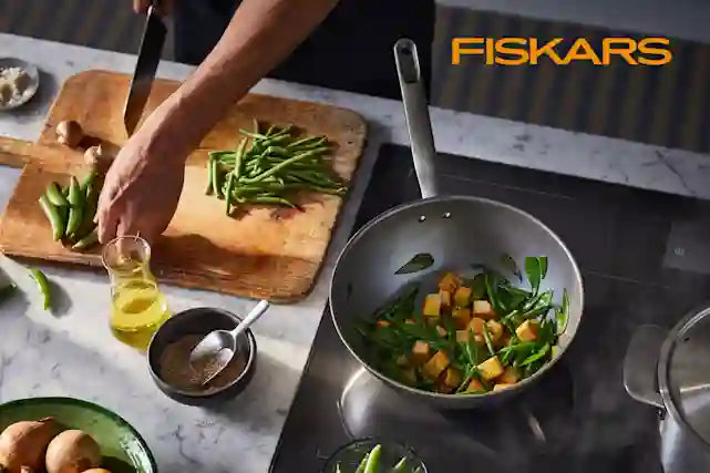 Oranssi Fiskars-logo oikeassa yläkulmassa. Kuvassa on keittiö, jossa tehdään ruokaa. Pilkotaan ja paistetaan juureksia.