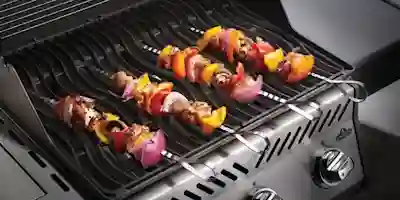 Tre färgglada spett med grönsaker och kött som grillas på en gasgrill.