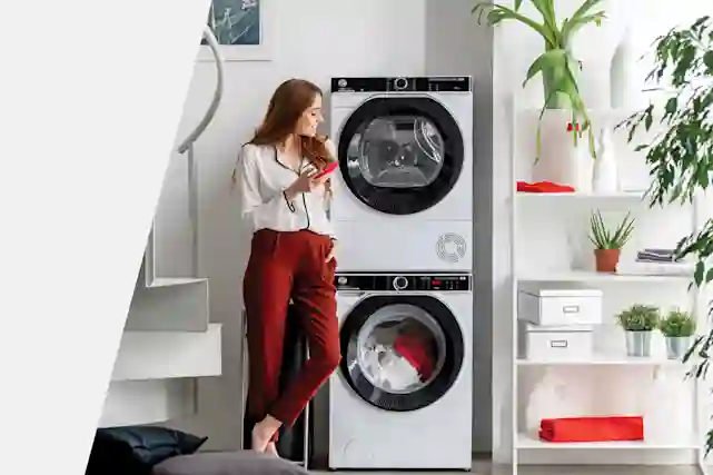 Henkilö nojaamassa Hooverin pesutorniin. Pesutornin toisella puolella hyllykkö , jossa viherkasveja ja pyyhkeitä.