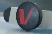Jääkiekko kaukalossa lähikuva kiekosta, joka on kääntynyt kyljelleen pystyasentoon. Kiekossa on punainen V-logo ja kiekon takana näkyy mailan lapa.