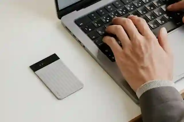 Hands typing on a laptop keyboard with a silver external hard drive beside it.