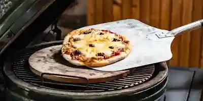 Pizzan lyfts med en metallpizzaspade från den runda pizzastenen på det gröna keramiska grillets galler.