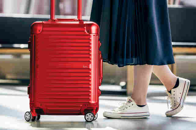 Punainen matkalaukku lentokentällä. Omistaja seisoo vieressä. Kuva on otettu niin läheltä, että näkyvissä on vain jalat.