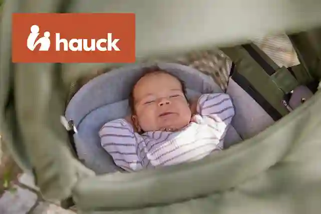 Vauva makaa hymyillen Hauck-merkkisessä vihreässä vaunussa, jossa on harmaa istuintyyny. Hauck-logo näkyy kuvassa.