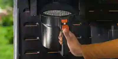 En hand öppnar en svart gasgrill vid det orangea handtaget. Inuti grillen syns brännaren och värmefördelare.
