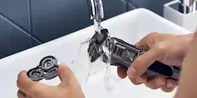 Hands rinsing the shaver head under running water in a white sink.