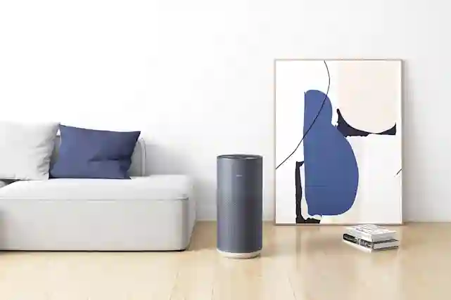 Ilmanpuhdistin huoneen lattialla, vasemmalla puolella harmaa sohva ja oikealla puolella kirjapino ja seinään nojaava taulu.