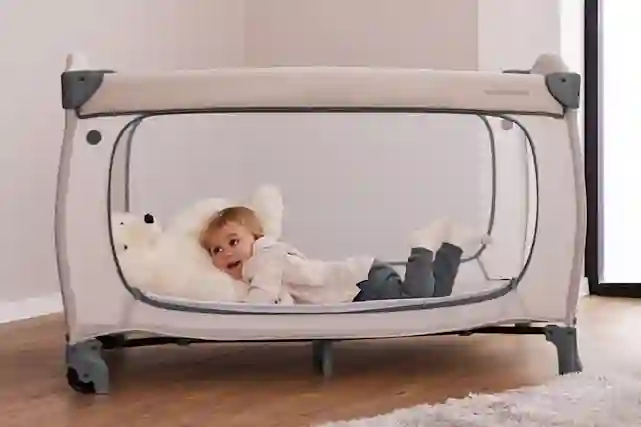 Hymyilevä lapsi makaa Sleep N Easyfold -matkasängyssä, jossa on valkoinen nallepehmolelu.