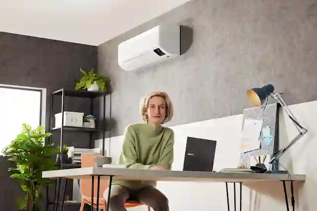 Nainen hymyilee työpöydällä tietokoneen ja kynien ääressä, seinällä ilmalämpöpumppu.