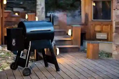 Kuva Traegerin pellettigrillistä grillaustarvikkeineen metsämaisemassa. Vieressä pöytä, jota vasten nojaa kalastusvälineet ja kirves. 