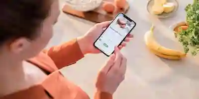 A person uses a smartphone in the kitchen to browse a recipe, surrounded by cooking ingredients and fresh vegetables.