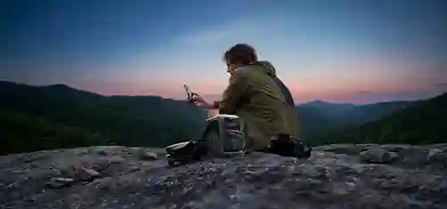 Henkilö istuu kallion päällä auringonlaskussa ja katsoo puhelintaan, vieressä kamera ja kannettava virtalähde.