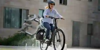 A smiling woman rides an electric bike through a fountain in an urban setting, wearing a helmet.