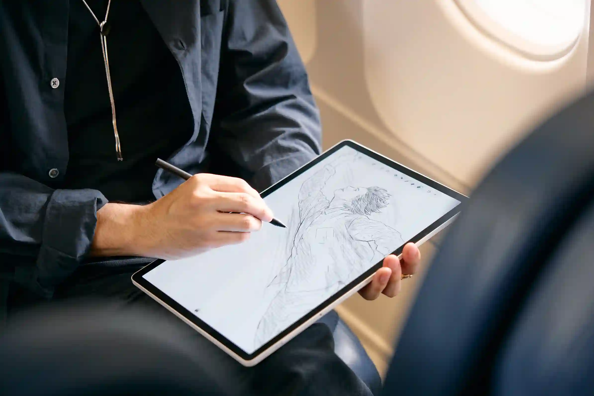 A person draws with a pen on a Wacom drawing tablet next to an airplane window.