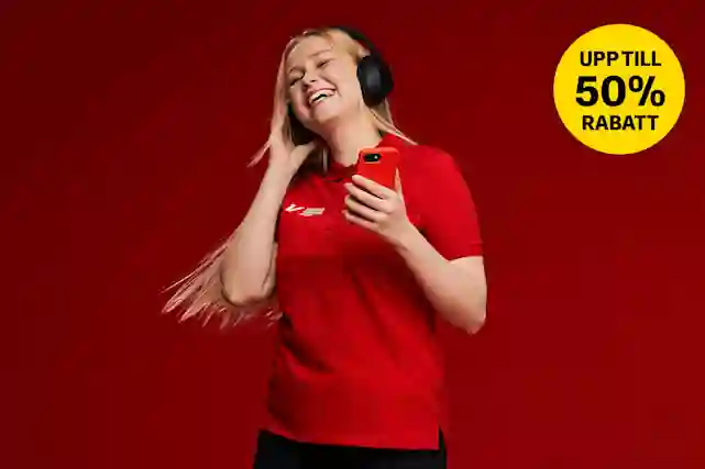 En leende kvinna i en röd Verkkokauppa.com-tröja lyssnar på musik med svarta hörlurar och håller en röd telefon.