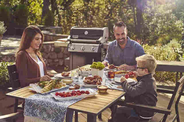 Perhe syö ulkona terassilla grilliruokaa pöydän ääressä aurinkoisena päivänä.