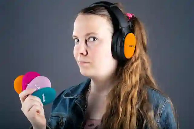 A woman wearing black headphones with orange Valco logos holds colorful Valco magnetic replacement covers.