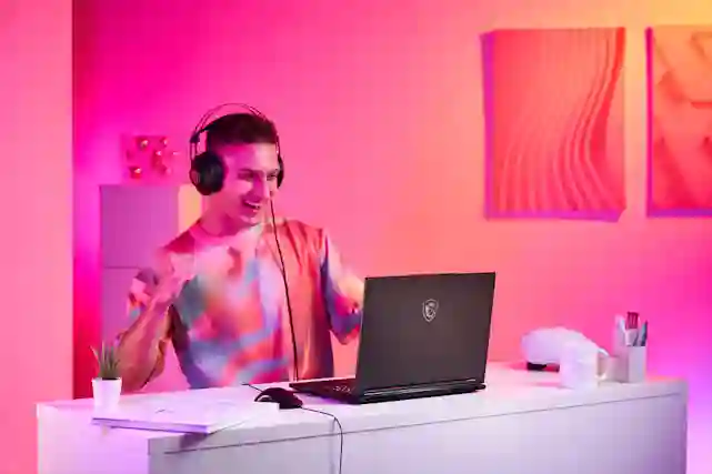 A smiling person wearing headphones plays on a laptop in pink and orange lighting.