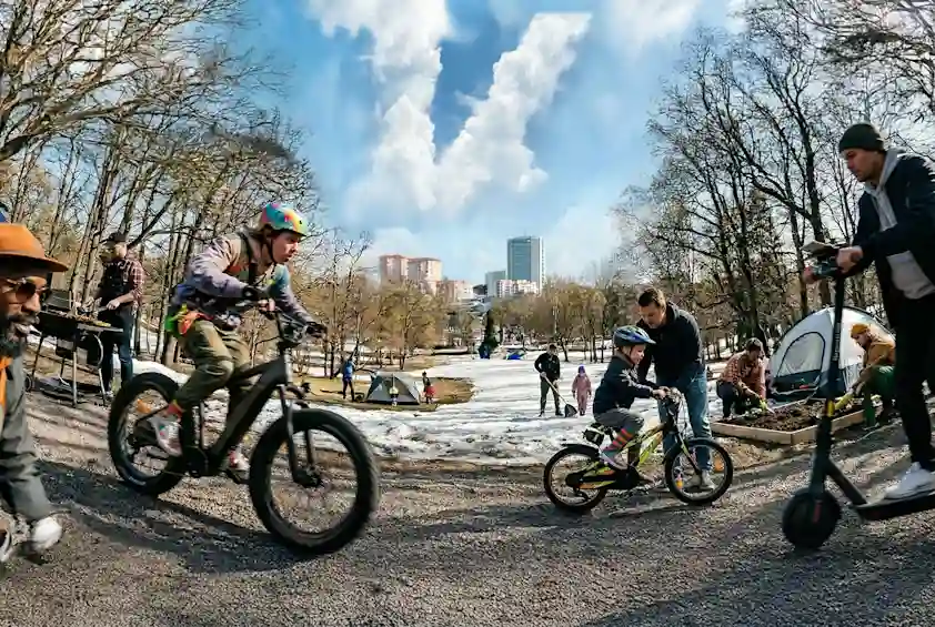 Keväinen puistomaisema, jossa lapset ja aikuiset pyöräilevät ja leikkivät ulkona. Etualalla sorapolku, taustalla puita ja kaupunkimaisema. Taivaalla V‑kirjaimen muotoinen pilvigrafiikka.
