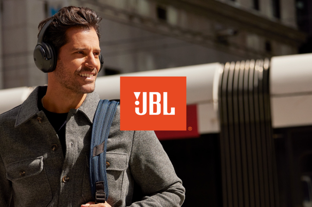 Henkilö kuuntelee musiikkia JBL Tour One M2 kuulokkeilla ja hymyilee. Taustalla kaupunki maisema. Edessä JBL-logo.
