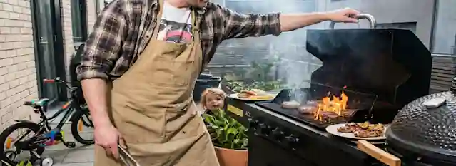 Kaasugrilli etupihalla käytössä. Grillissä näkyy pienet liekit ja paistuvia pihvejä. Vieressä tarjolle nostettua ruokaa ja Anton Oliverin Kamado-hiiligrilli.