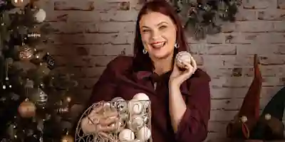A smiling woman holds a Christmas ornament in front of a brick wall, with a Christmas tree and decorations around her.