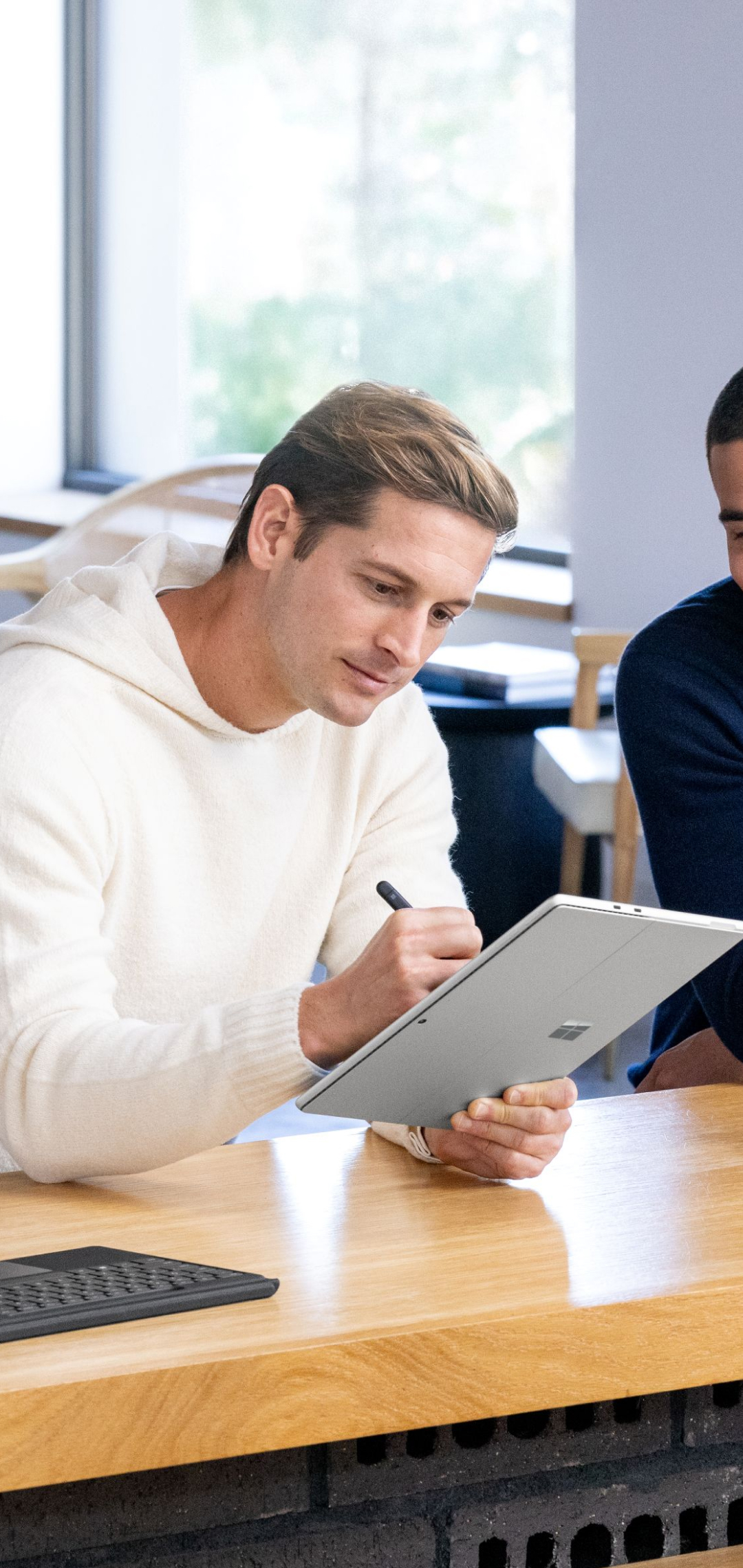 A man working on a Surface Pro using a digital pen and a detachable keyboard at a table next to a coworker