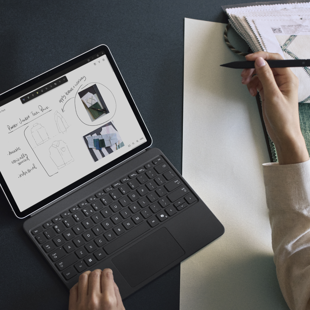 A top-down view of a woman sketching on a Surface Pro device with a Surface Pen while at a table covered in fabric and paper