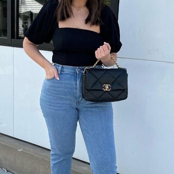 a woman in denim pants and a black top holding a black Chanel 19