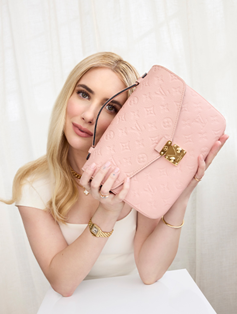 Emma Roberts wearing a short sleeve white dress and holding up a light pink monogram Louis Vuitton Empreinte Pochette Metis bag close to her face