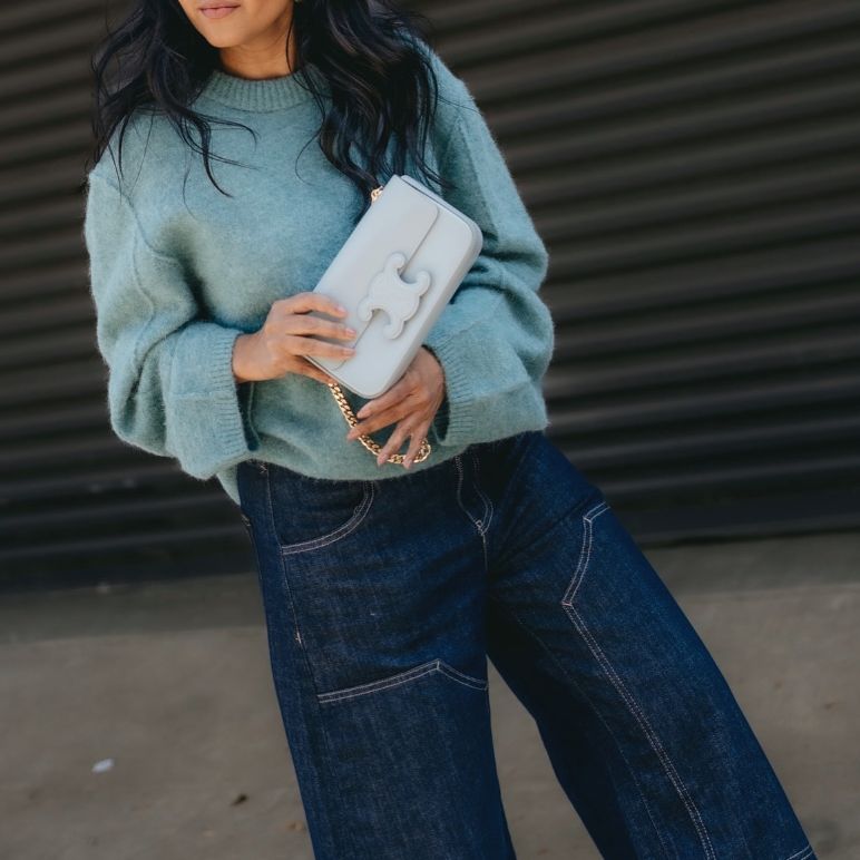 a woman in denim pants and a light blue sweater holding a CELINE Shiny Calfskin Cuir Triomphe Chain Shoulder Bag in Ice Blue