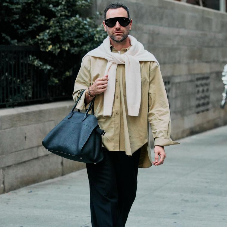 a man in a beige long sleeve top and cream sweater with black pants holding a dark The Row Margaux